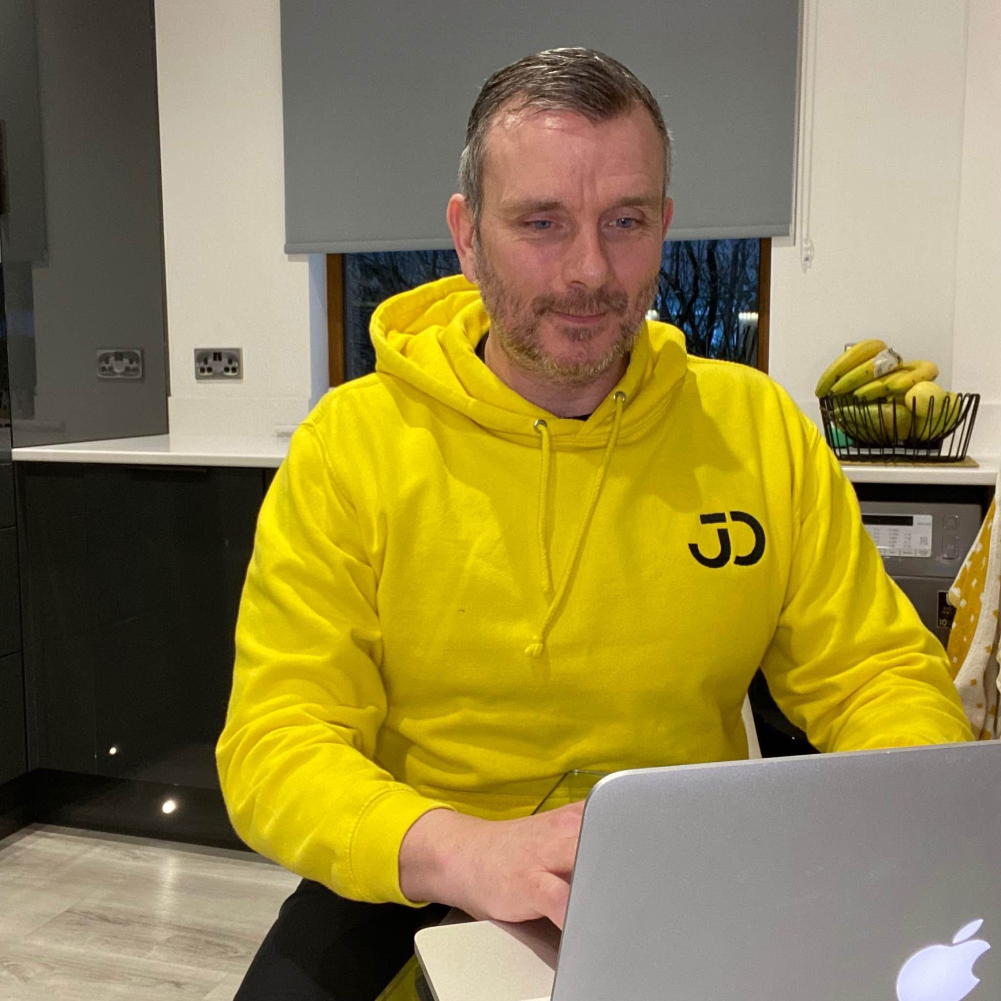 A man wearing a yellow hoodie working on a laptop.